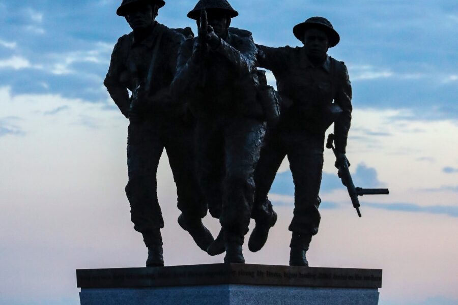 A statute representing three British soldiers stands in front of a dull sunset.
