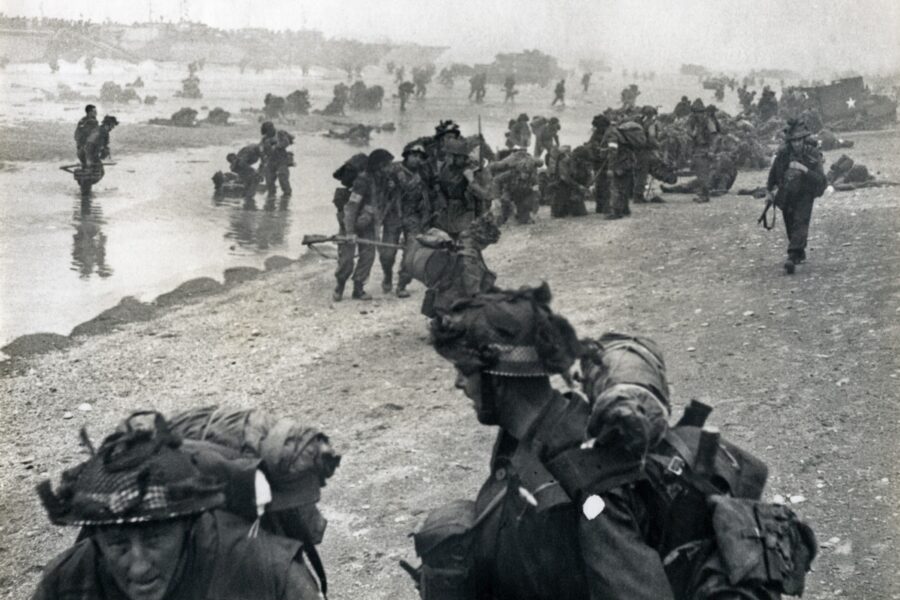Soldiers form a line on a Normandy beach while in the background their comrades run through the sea towards them.