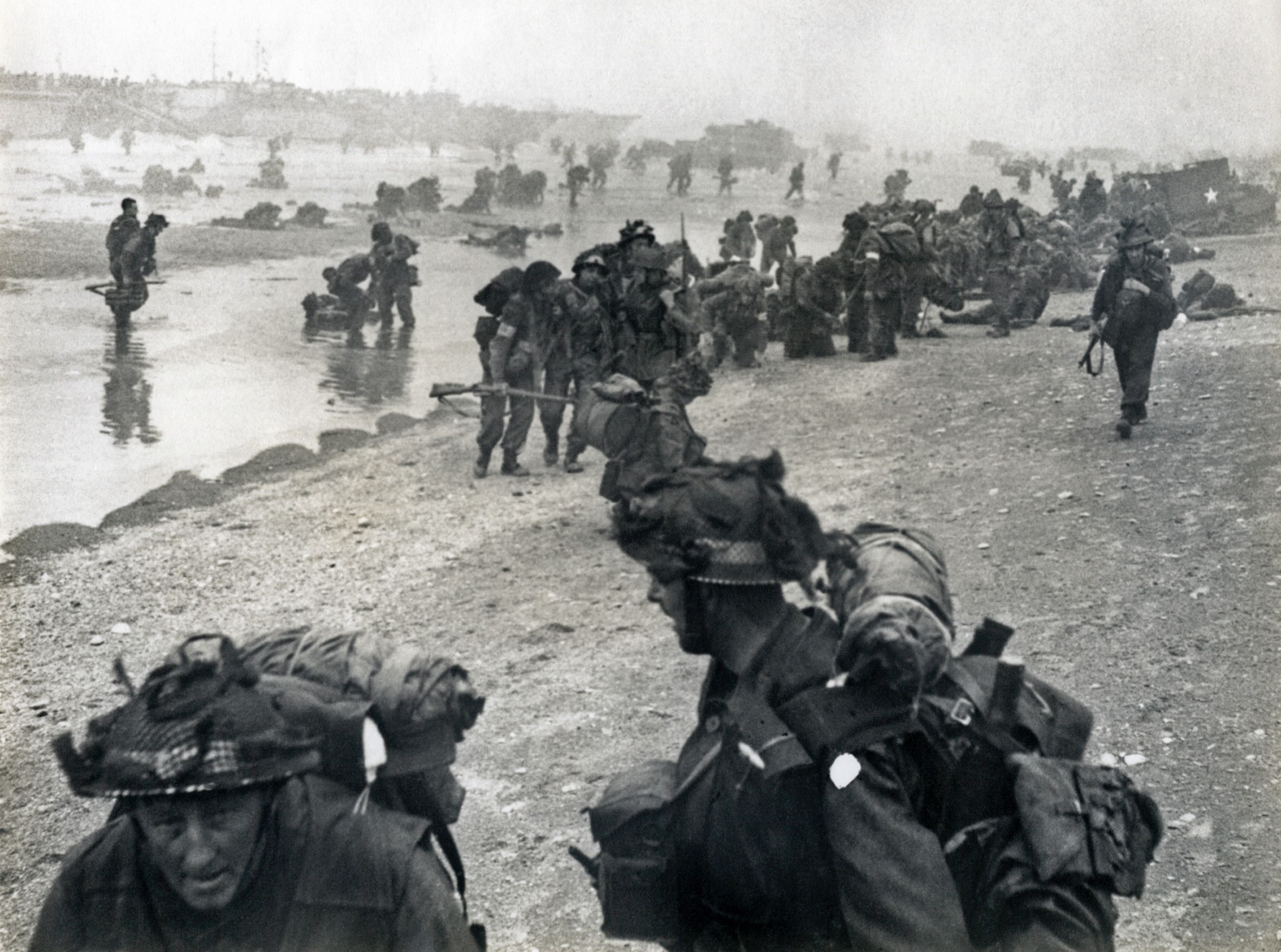 Soldiers form a line on a Normandy beach while in the background their comrades run through the sea towards them.