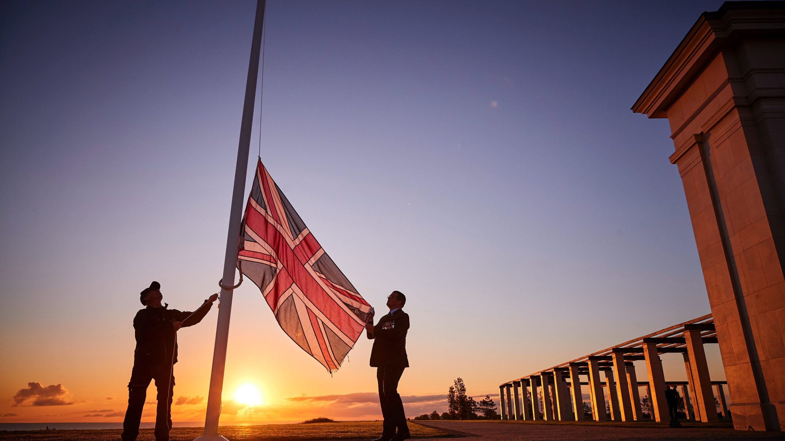 Two silhouetted figures raise a Union flag in front of a bright sunrise. The stone pillars of the British Normandy Memorial stand to the right.
