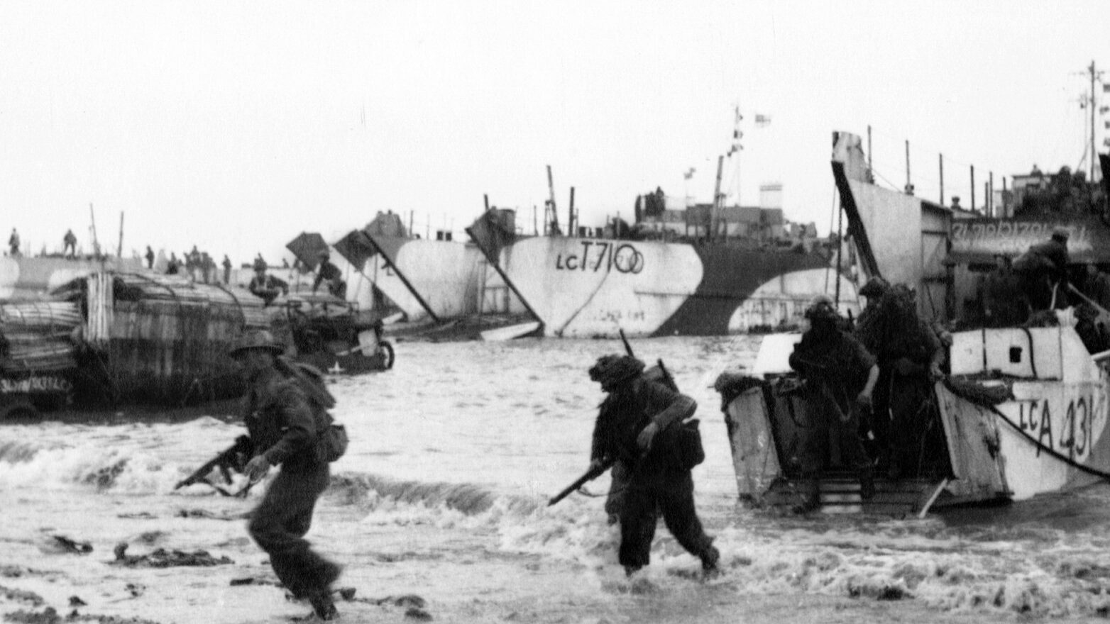 Soldiers leap from a landing craft into the shallow water and run onto the beach.