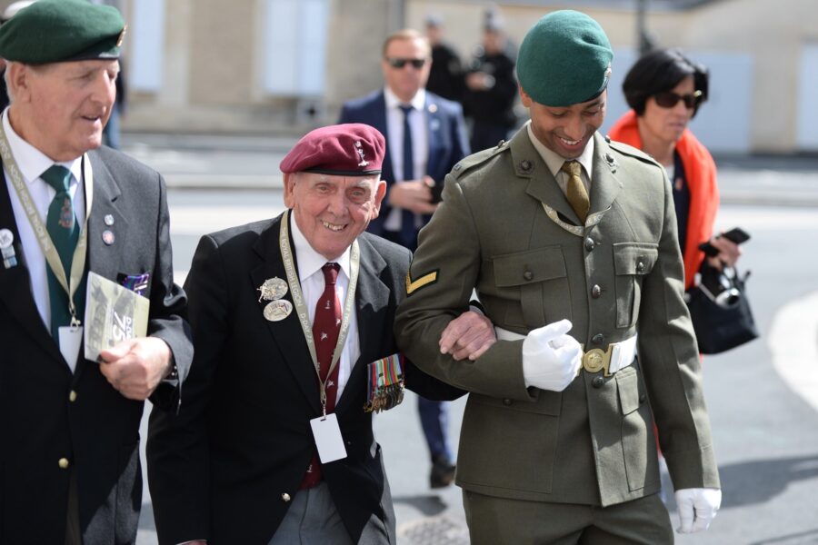 An elderly veteran wearing a suit and beret smiles as he links arms with a soldier in dress uniform.